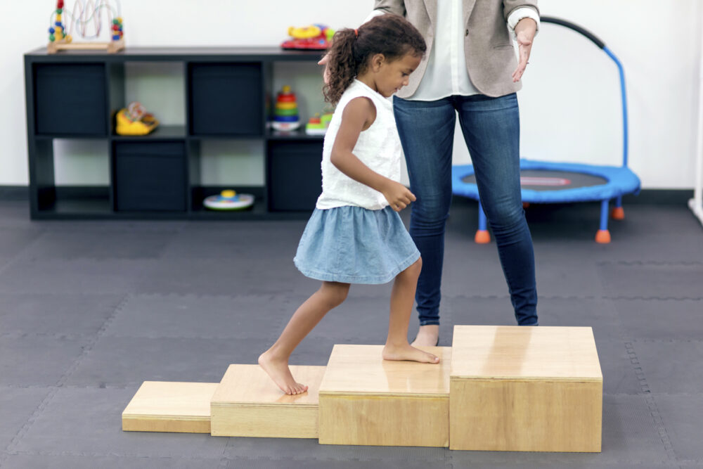 petite fille apprenant à monter des marches sur des caisses en bois en séance de thérapie intensive appelée "Dynamic Movement Intervention".